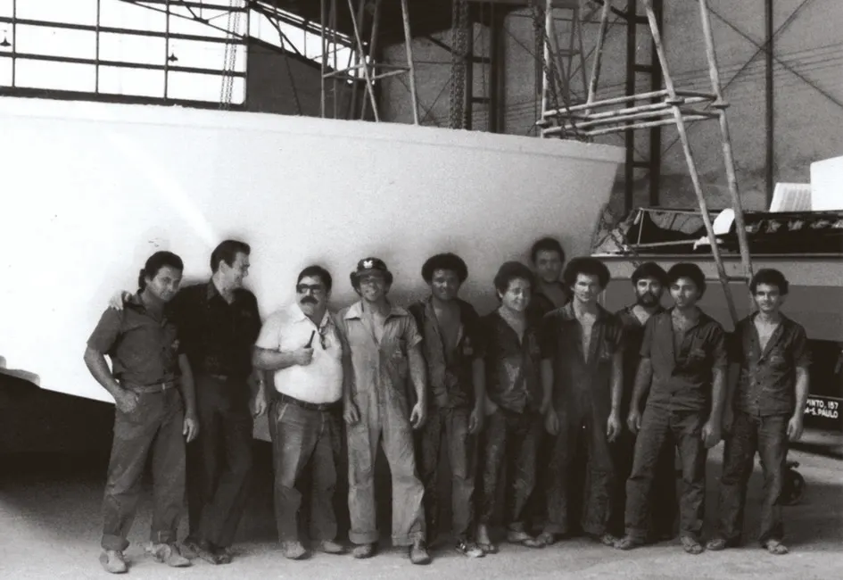 A equipe do estaleiro nos primeiros anos, enfileirada em frente ao casco cru de um barco em construção